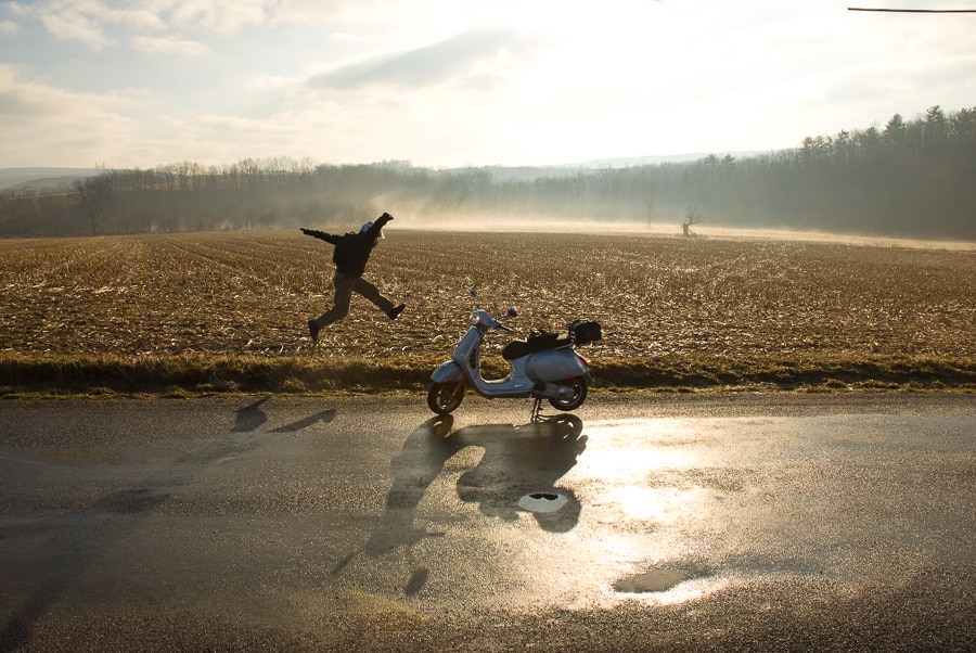 Man jumping in air next to a Vespa scooter at sunrise
