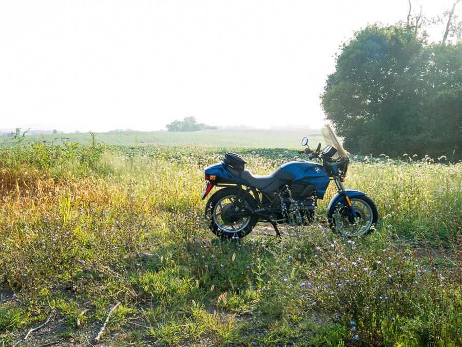 BMW K75 motorcycle parked along a farm field in dazzling morning sunlight.