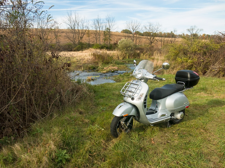 Vespa scooter parked in a field next to a creek.