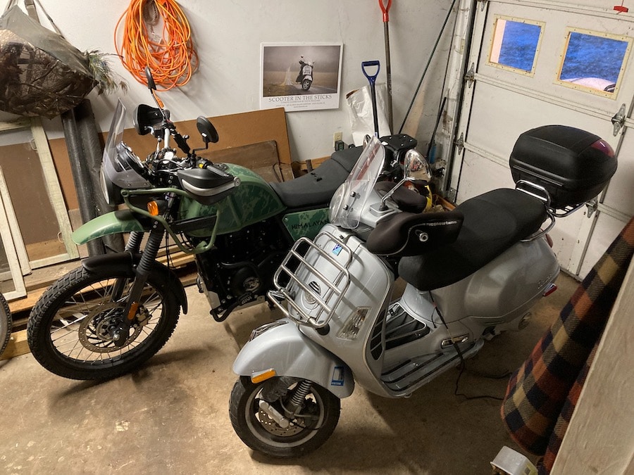 Royal Enfield Himalayan motorcycle and Vespa GTS scooter in a garage.