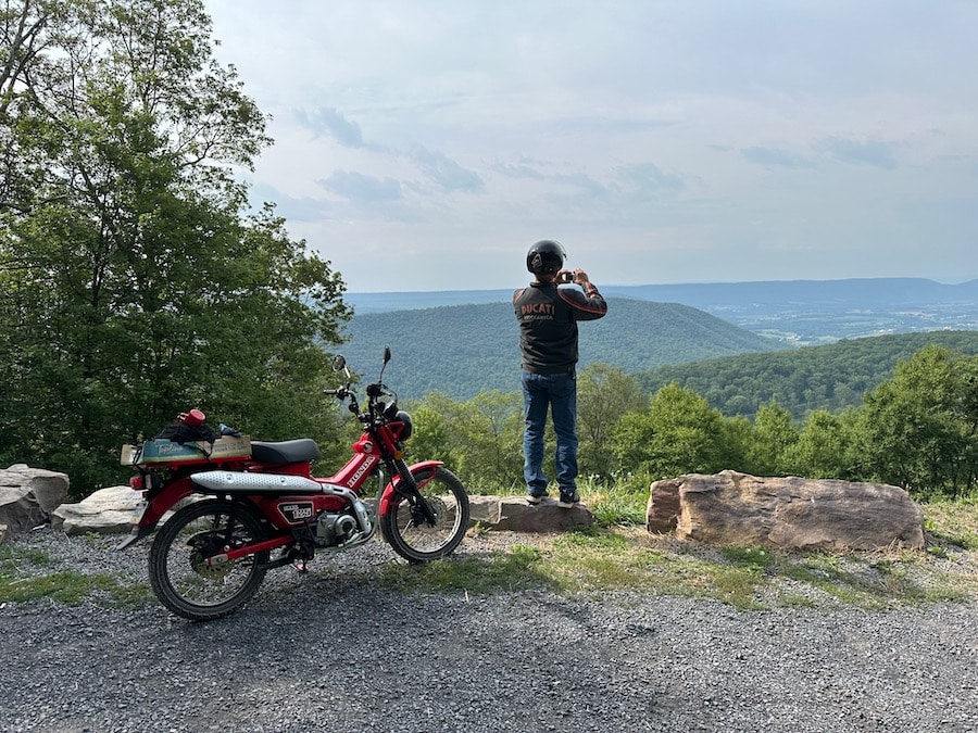 Man at scenic overlook with a Honda Trail 125 motorcycle.