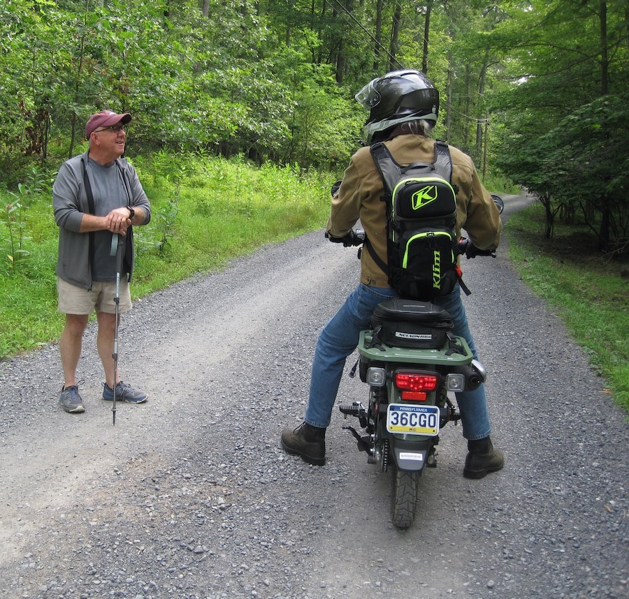 Man on a motorcycle talking to a pedestrian on a gravel road.