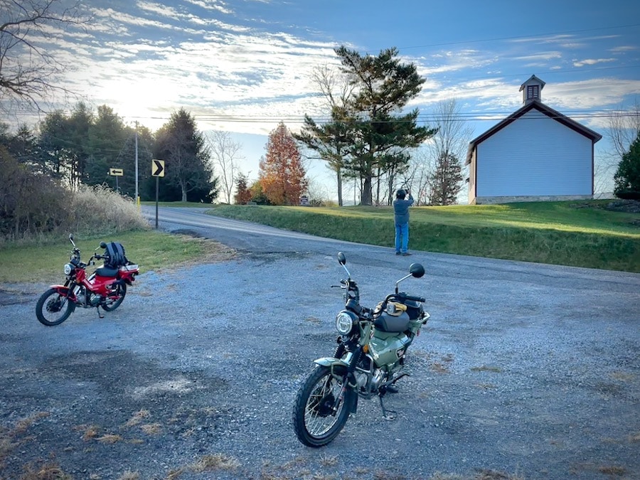 Two Honda Trail 125 motorcycles parked on a gravel lot by an old one-room school.