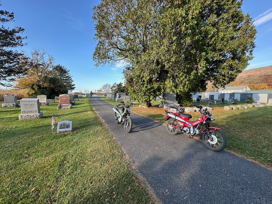 Two Honda Trail 125 motorcycles parked at a cemetery.