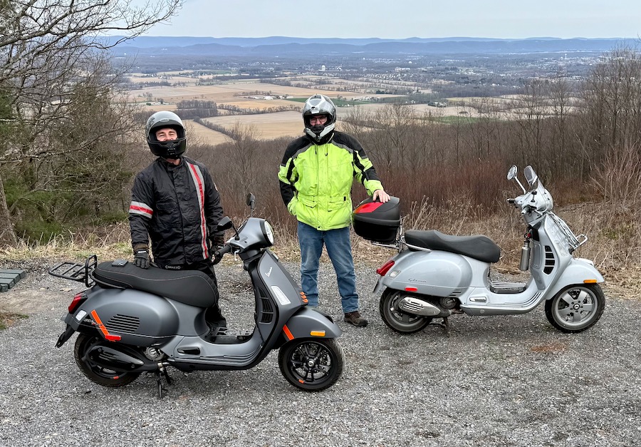 Two Vespa scooter riders stopped at a scenic vista.