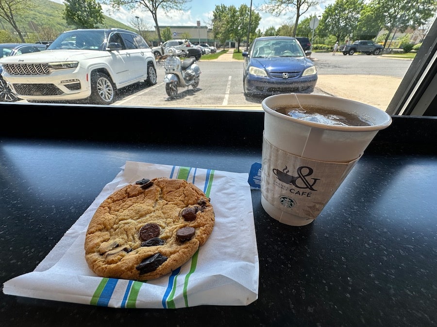 A chocolate chip cookie and cup of tea on a counter by a window with a Vespa scooter parked outside.