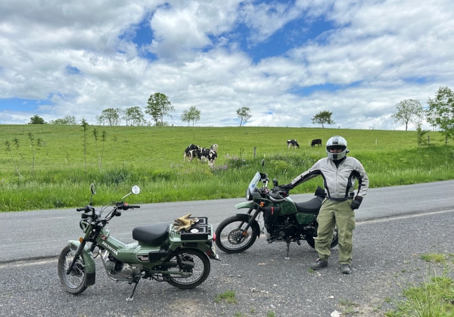 A motorcycle rider standing along a rural road with a Royal Enfield Himalayan and Honda Trail 125 motorcycle.