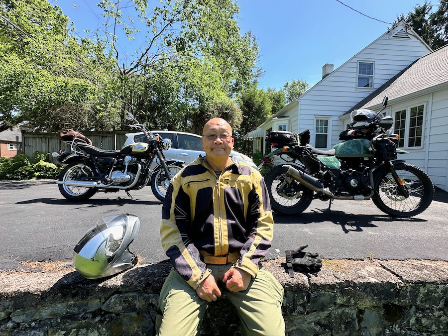Man sititng on a wall with two motorcycles behind him.