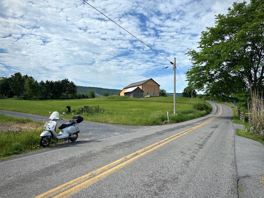 Vespa GTS scooter on a rural road with a barn in the background.