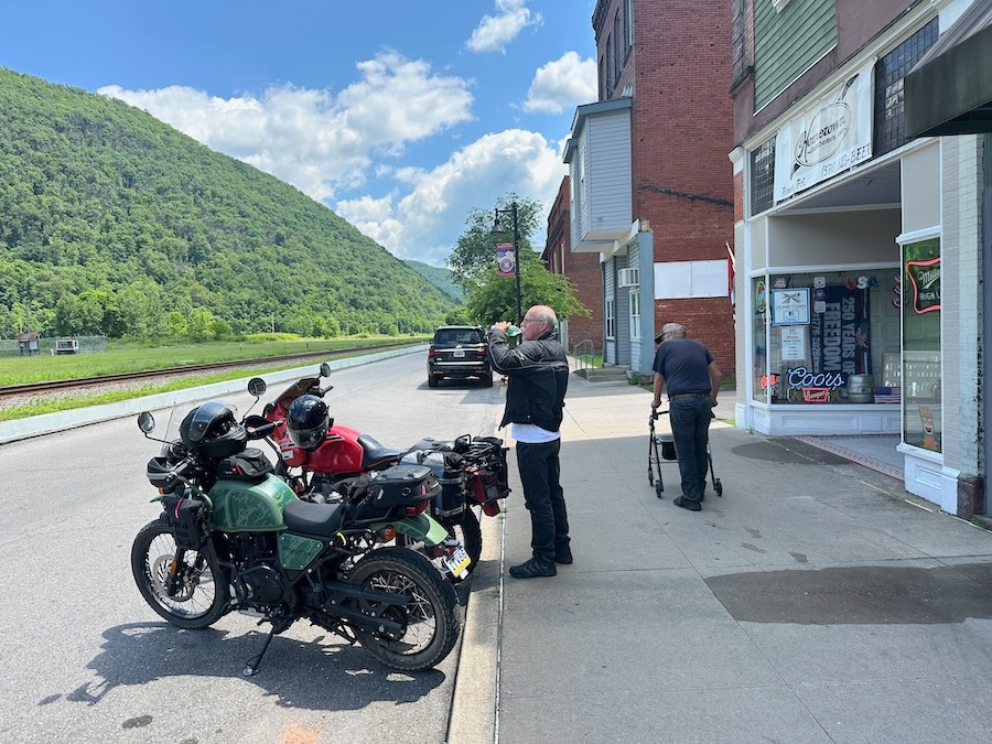 Man standing on a sidewalk drinking water by two motorcycles.