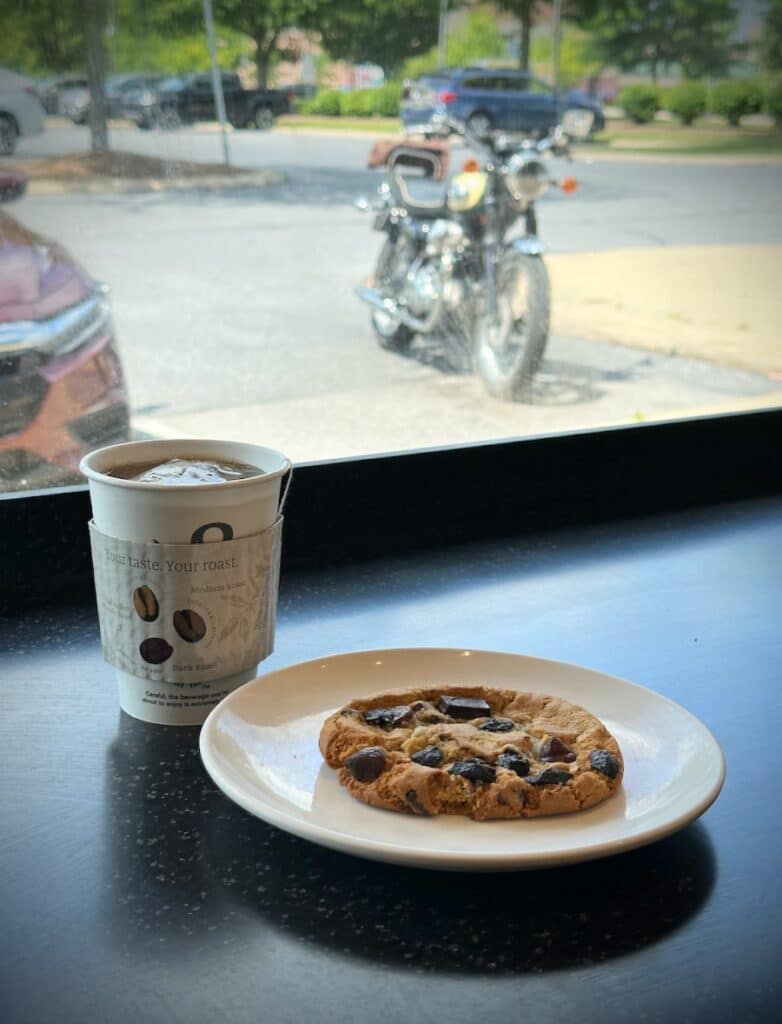 A hot tea and big chocolate chip cookie sitting on a counter with a motorcycle parked outside the window.