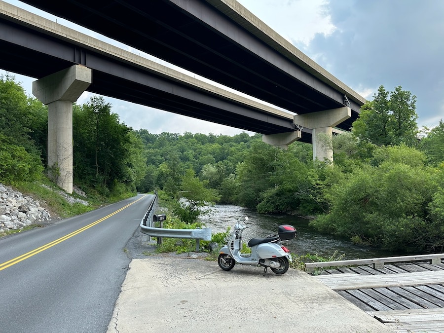 A 2006 Vespa GTS 250 scooter parked along a rural road under an Interstate highway bridge.