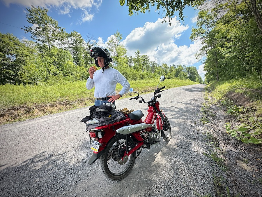 Paul Ruby standing next to his Honda Trail 125 motorcycle eating lunch.