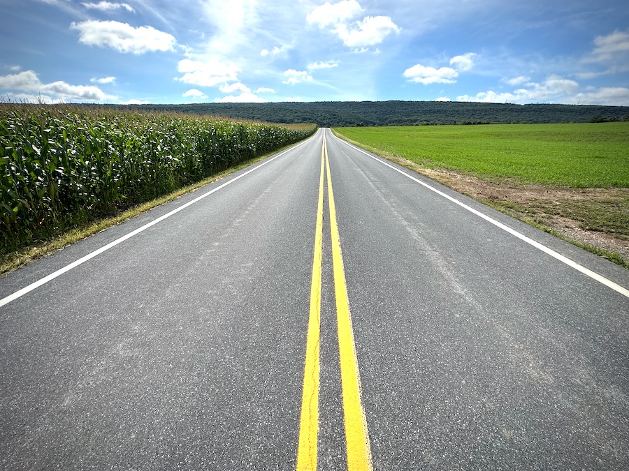 An empty, straight two-lane road on a sunny day with two bright yellow lines.