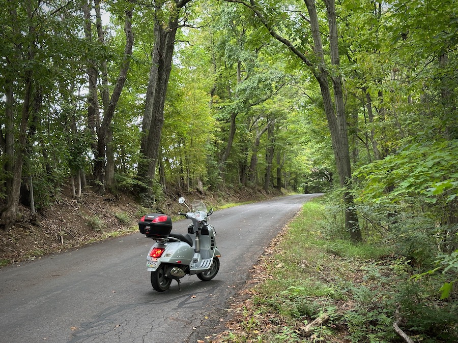A Vespa GTS scooter on a tree-lined rural road.