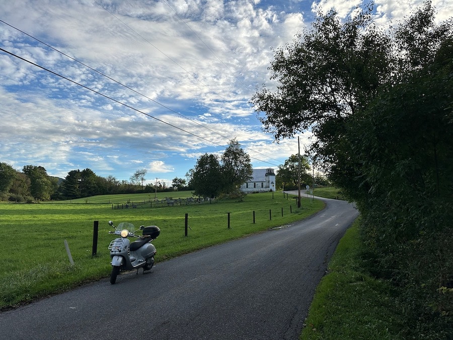 A Vespa GTS scooter parked along a rural road with an old church in the background.