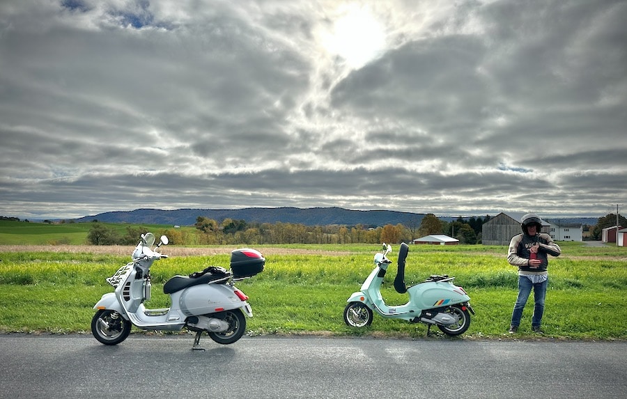 Two Vespa scooters parked along a rural road.