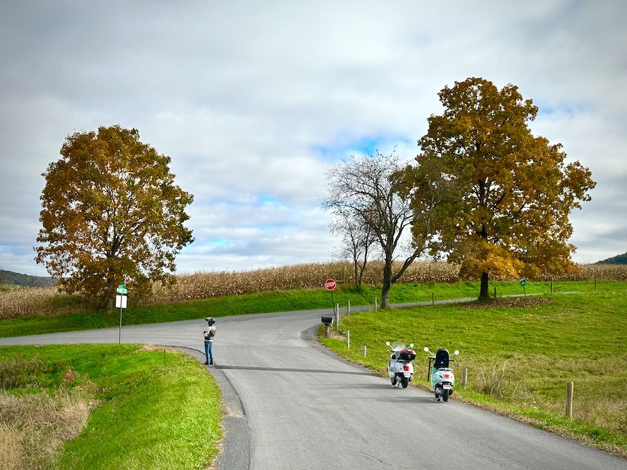Two Vespa scooters parked at a rural intersection while a man shoots pictures.